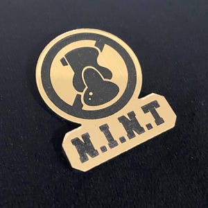 May include: A gold-colored pin with a black circular design featuring a stylized figure. Below the circle is a gold rectangle with the letters "N.I.N.T." in black block letters. The pin is likely a collectible or accessory.