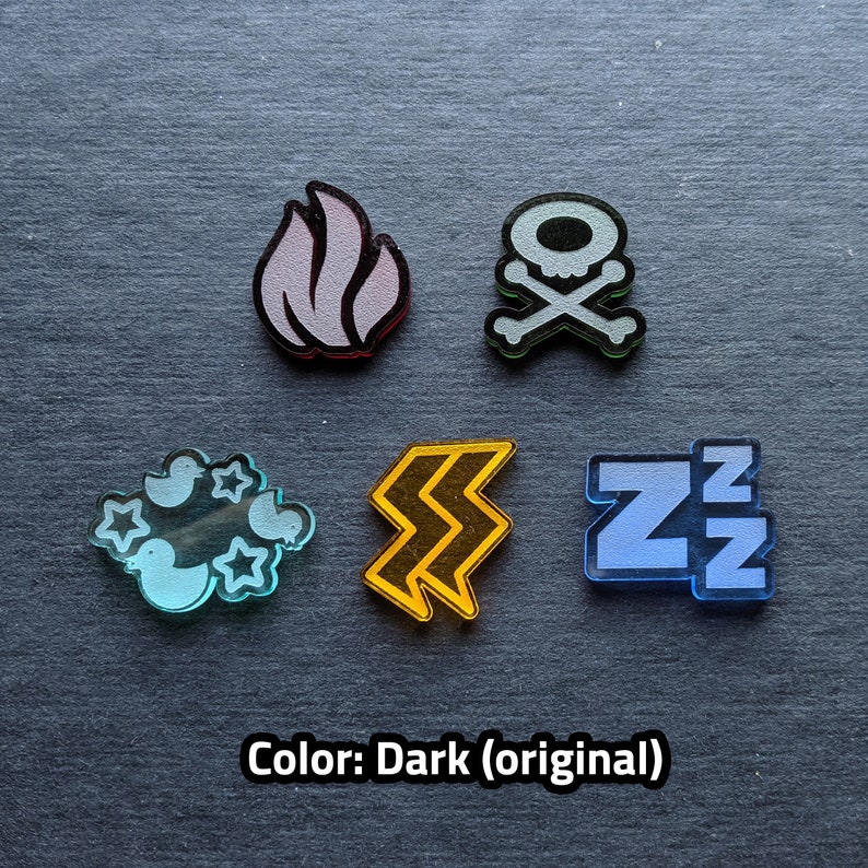 Puede incluir: Cinco fichas acr&iacute;licas de diferentes colores con varios s&iacute;mbolos. Las fichas son una llama rosa claro, una calavera y huesos cruzados grises, una nube azul claro con p&aacute;jaros, un rayo amarillo y un s&iacute;mbolo 'ZZ' azul. Las fichas est&aacute;n sobre un fondo gris oscuro. Color: Oscuro (original)