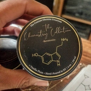 May include: A black and gold label with the text "The Couture Chemist Creations The Chemistry Collection -Serotonin- NH2 ~Vanilla Plum ~ Hand-Poured in NY | 6 oz. | 178 ml" and a chemical structure diagram.