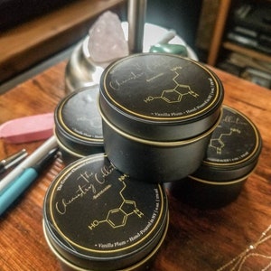May include: Three black candle jars with gold lids and labels. The labels feature a chemical formula and the text "The Chemistry Collection" and "Serotonin". The jars are stacked on top of each other on a wooden surface.