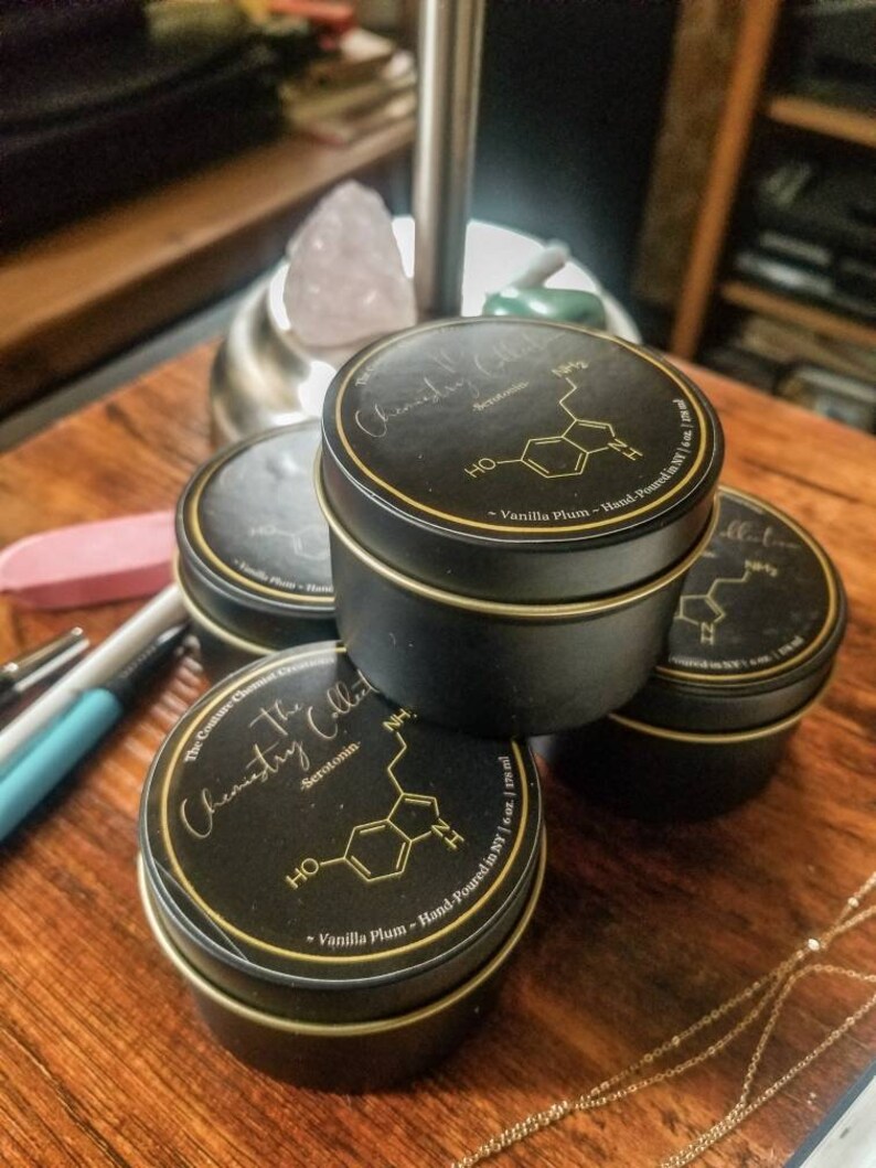 May include: Three black candle jars with gold lids and labels. The labels feature a chemical formula and the text "The Chemistry Collection" and "Serotonin". The jars are stacked on top of each other on a wooden surface.