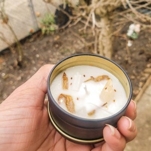 May include: A small, white candle with gold flecks and crystals in a black and gold tin.