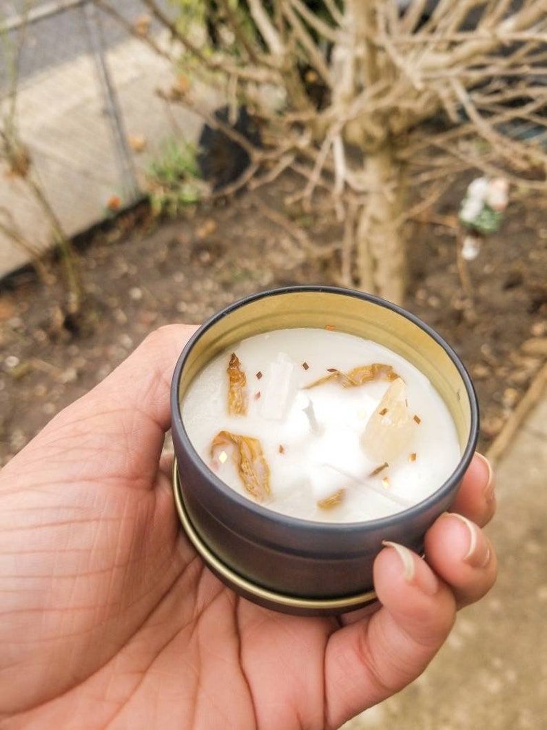 May include: A small, white candle with gold flecks and crystals in a black and gold tin.