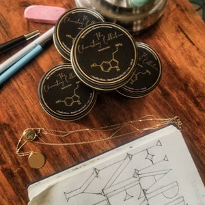 May include: Four black round containers with gold lettering that reads "The Chemistry Collection" and a chemical formula. The containers are on a wooden surface with a gold chain necklace and a notebook with the word "HAPPY" drawn in black ink.