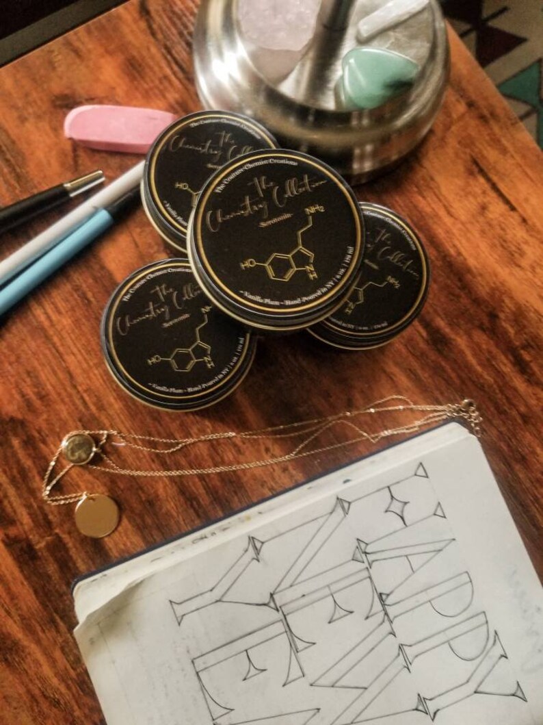 May include: Four black round containers with gold lettering that reads "The Chemistry Collection" and a chemical formula. The containers are on a wooden surface with a gold chain necklace and a notebook with the word "HAPPY" drawn in black ink.