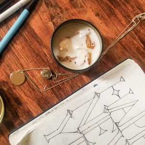 May include: A white candle in a clear glass jar with a gold lid. The candle has a natural, earthy scent and is decorated with dried flowers and spices. The candle is sitting on a wooden table with a notebook and pens.