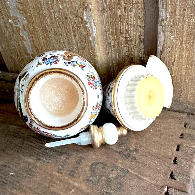 May include: A vintage, open perfume bottle with a decorative design. The bottle is white with colorful floral and bird motifs, accented with gold trim. The lid and pump are separate, showing the interior.