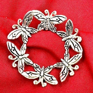May include: A silver-toned metal ring with a design of multiple butterflies surrounding a central open space. The ring is on a red fabric background.