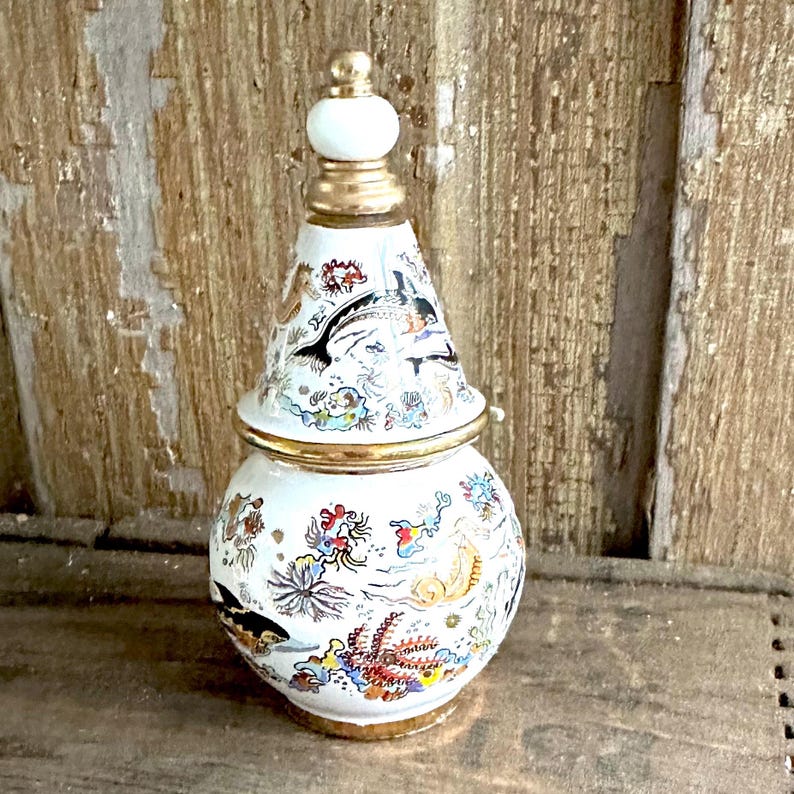 May include: A white ceramic jar with a conical lid, adorned with marine life illustrations in various colors. The jar has gold trim and a white finial. The illustrations include fish, coral, and other sea creatures. The jar is approximately 6 inches tall.