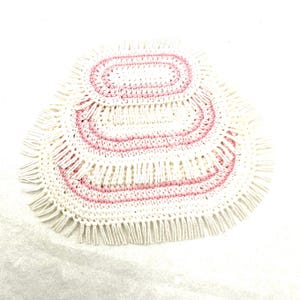 May include: A handmade, oval-shaped, off-white crochet doily with pink accents. The doily features concentric oval patterns in pink, with a fringe border. The craftwork is visible, showcasing the texture and detail of the yarn.