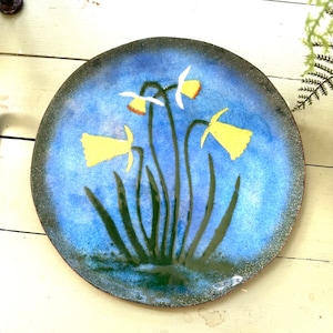 以下が含まれることがあります： 青い背景と花の模様が施された丸い装飾皿。 黄色い水仙と緑の茎と葉が特徴です。 皿の縁は濃い緑色です。