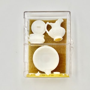 Könnte beinhalten: Ein Set aus Miniatur-Küchenutensilien aus weißem Porzellan in einer durchsichtigen Plastikbox mit braunem Filzbezug. Das Set enthält eine Kanne, eine Schüssel, eine abgedeckte Schale und einen kleinen Topf.