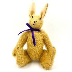 Op de afbeelding: Een pluche, lichtbruin konijn met lange oren en een paars lint om zijn nek.