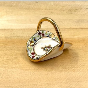 May include: A small white ceramic basket with a gold handle and floral design. The basket is decorated with red, blue, and yellow flowers.