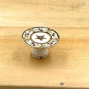 May include: A small white ceramic cake stand with a gold rim and floral design. The top of the stand has a white plate with a gold rim and a floral design.