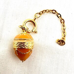 May include: A gold-toned acorn-shaped pendant with a chain. The pendant features a two-tone orange and cream enamel design, accented with gold-colored metal detailing. A gold chain is attached to the pendant with a clasp.