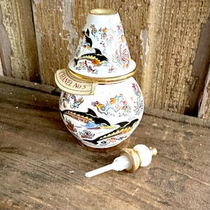 May include: A vintage white perfume bottle with a decorative lid, featuring a design of black and yellow fish. The bottle has gold accents and the text "CHANEL No 5". The bottle's stopper is white and gold.