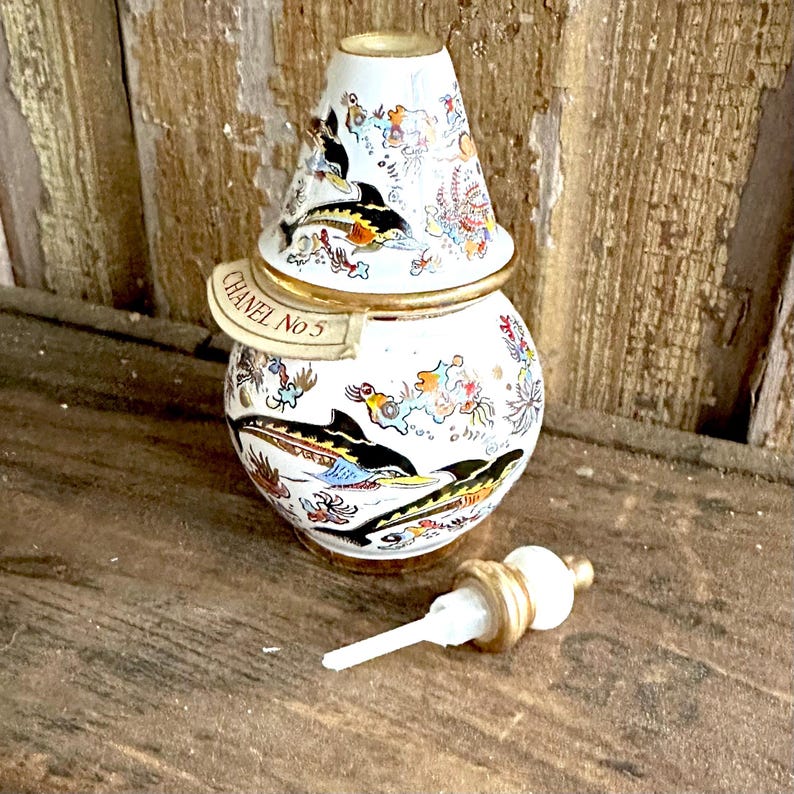 May include: A vintage white perfume bottle with a decorative lid, featuring a design of black and yellow fish. The bottle has gold accents and the text "CHANEL No 5". The bottle's stopper is white and gold.