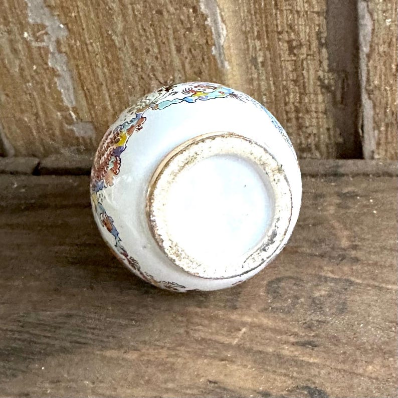 May include: A small, white ceramic vase with a rounded shape. The vase features colorful floral patterns in shades of red, blue, and yellow. The rim has a gold-colored trim, and the base is unglazed.