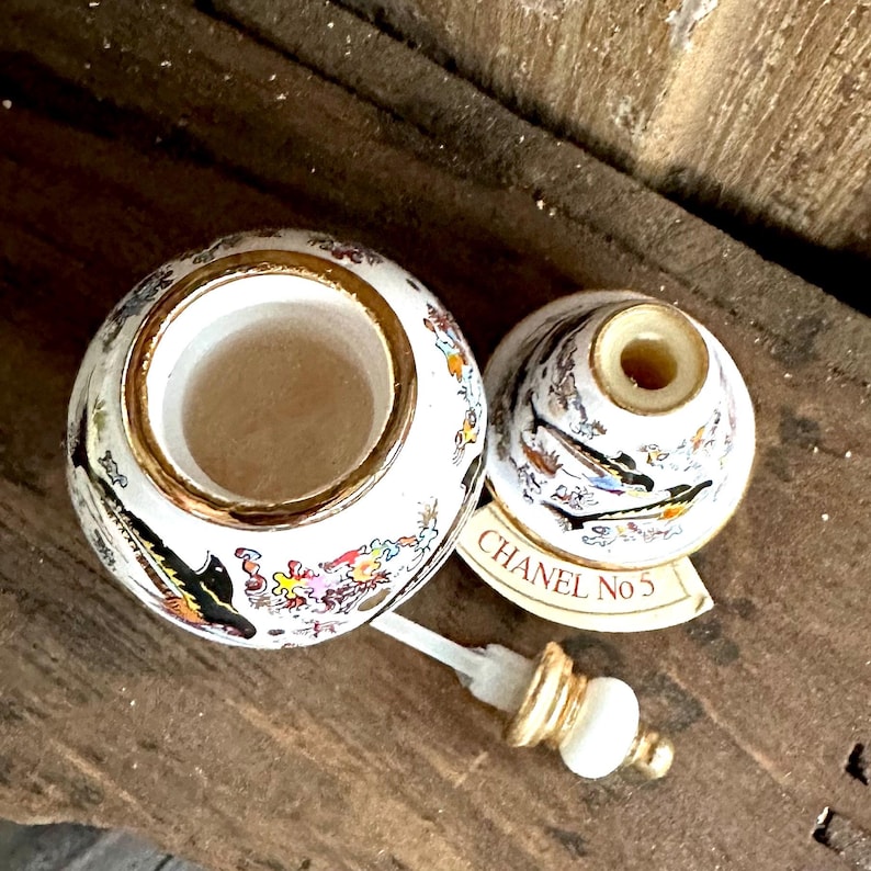 May include: A vintage white and gold perfume bottle with a decorative design. The bottle is open, revealing the interior, and the lid is nearby. The bottle has a gold rim and a decorative design with birds and flowers. The label reads "CHANEL No 5".