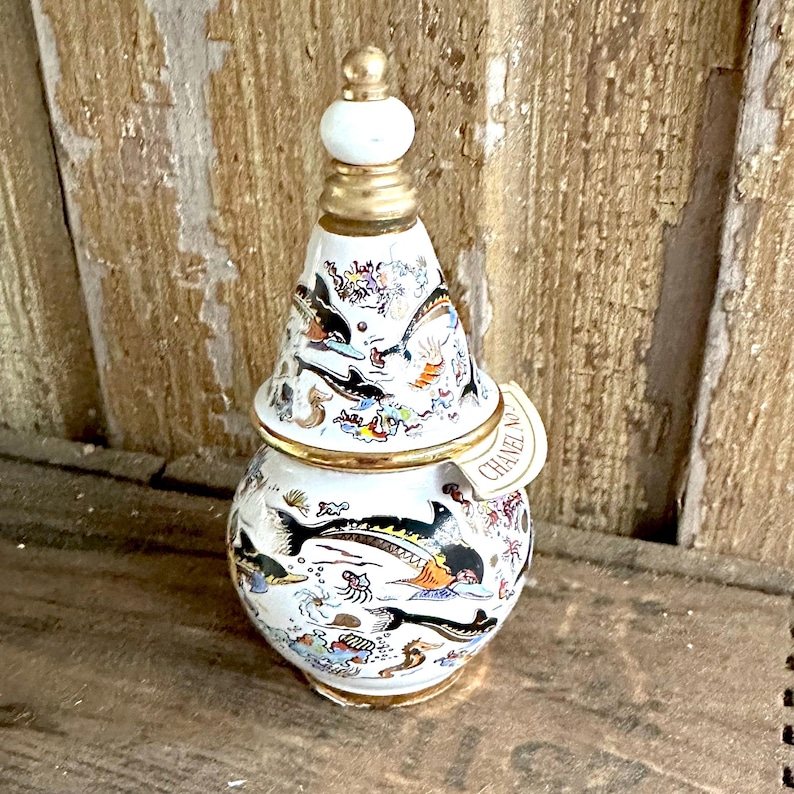 May include: A decorative white ceramic jar with a lid, featuring a marine life design with black, orange, and blue accents. The jar has gold trim and a white finial. A tag reads "CHANEL No."