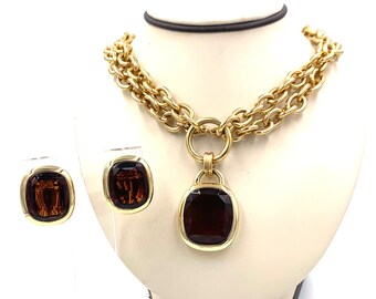 May include: A gold-toned necklace and matching earrings set. The necklace features a double-strand chain with a large pendant and a pair of square earrings. The pendant and earrings have a dark brown, square-cut stone.