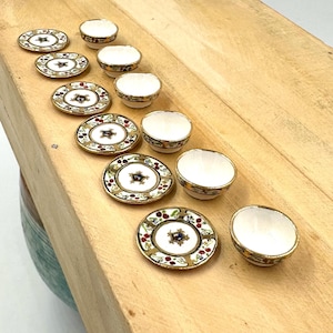May include: Set of ten white ceramic teacups and saucers with gold trim and colorful floral designs. The cups and saucers are arranged in a row on a wooden surface.