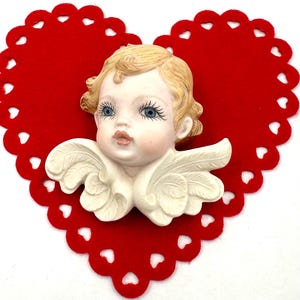 May include: A porcelain cherub figurine with blonde hair and blue eyes, wearing white wings. The figurine is set against a red felt heart with a scalloped edge.
