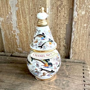 May include: A decorative white perfume bottle with a gold-colored rim and stopper. The bottle is adorned with colorful illustrations of fish and other sea creatures. The text "CHANEL No 5" is displayed on the bottle.