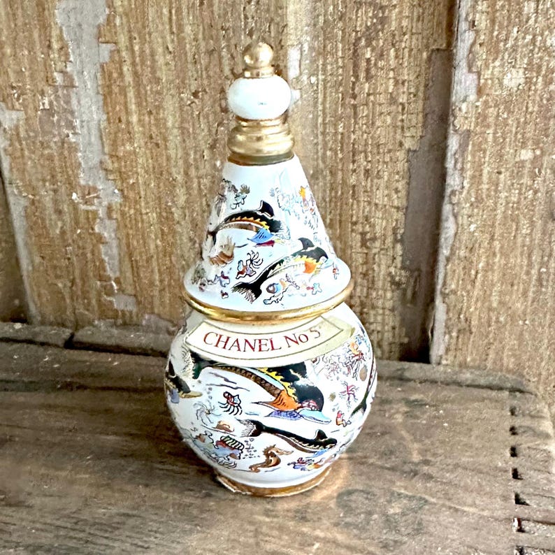 May include: A decorative white perfume bottle with a gold-colored rim and stopper. The bottle is adorned with colorful illustrations of fish and other sea creatures. The text "CHANEL No 5" is displayed on the bottle.