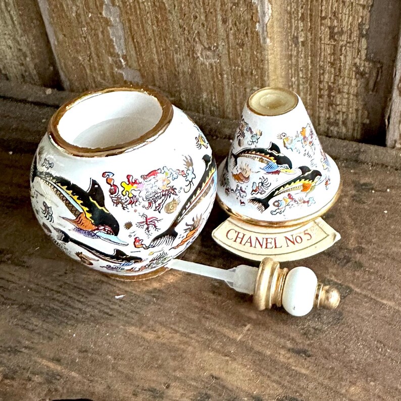 May include: A white ceramic perfume bottle and stopper set with gold trim. The bottle and lid are decorated with black, yellow, and orange sea creature illustrations. The label reads "CHANEL No 5".