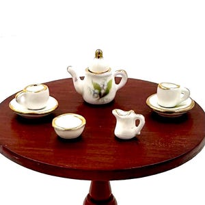 May include: A miniature porcelain tea set on a wooden table. The set includes a teapot, creamer, sugar bowl, and two teacups and saucers. The teapot has a floral design and gold accents.