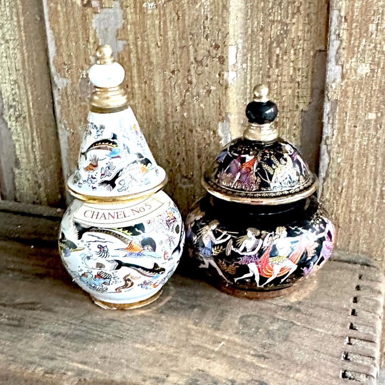 May include: Two decorative ceramic perfume bottles. One is white with a conical lid, gold trim, and the text "CHANEL No 5". The other is black with a rounded lid and gold accents. Both bottles feature detailed painted designs.