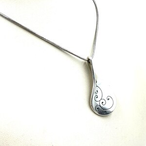 以下が含まれることがあります： 滑らかなティアドロップ型のチャームと光沢のある仕上げが特徴のシルバー調ペンダントネックレス。ペンダントの表面には複雑で暗い渦巻き模様が刻まれており、細いスネークチェーンから吊り下げられています。