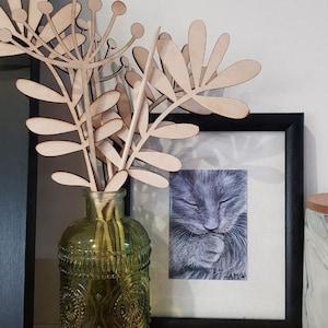 May include: An arrangement featuring a green glass vase with embossed detailing, holding wooden floral stems. A framed grayscale cat portrait is positioned behind the vase. A marble jar with a wooden lid is on the right.