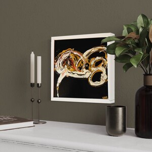 Puede incluir: Una pintura abstracta enmarcada en blanco con remolinos dorados y blancos sobre un fondo negro. La pintura est&aacute; colgada en una pared marr&oacute;n sobre un estante blanco con un libro, un candelero y un jarr&oacute;n con hojas verdes.