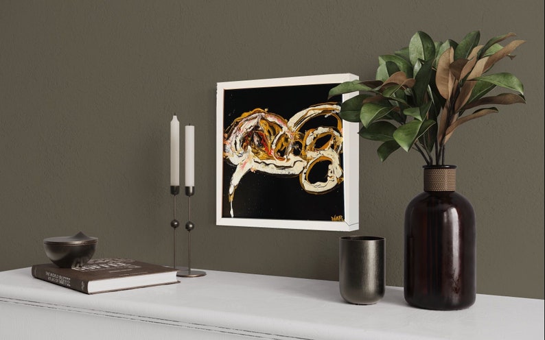 Puede incluir: Una pintura abstracta enmarcada en blanco con remolinos dorados y blancos sobre un fondo negro. La pintura est&aacute; colgada en una pared marr&oacute;n sobre un estante blanco con un libro, un candelero y un jarr&oacute;n con hojas verdes.