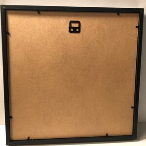 Puede incluir: Marco de imagen negro con un respaldo de cart&oacute;n marr&oacute;n. El marco tiene un gancho de metal para colgar en la parte posterior.