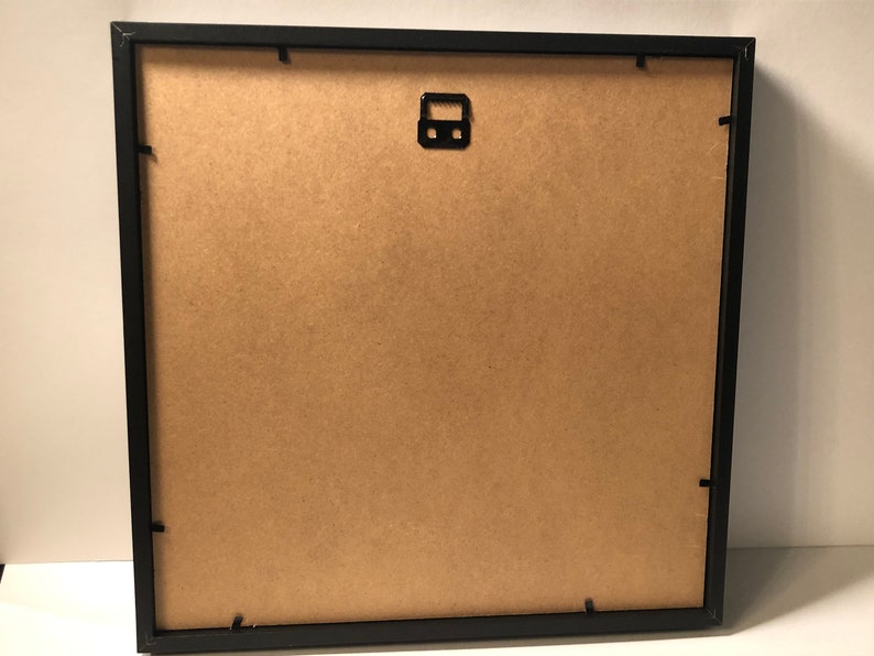 Puede incluir: Marco de imagen negro con un respaldo de cart&oacute;n marr&oacute;n. El marco tiene un gancho de metal para colgar en la parte posterior.