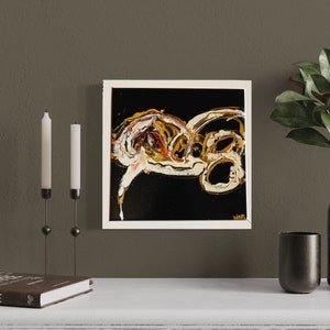 Puede incluir: Pintura abstracta en un marco blanco con pintura dorada, blanca y negra sobre fondo negro. La pintura est&aacute; colgada en una pared marr&oacute;n sobre una repisa blanca con un libro, velas y un jarr&oacute;n con hojas verdes.