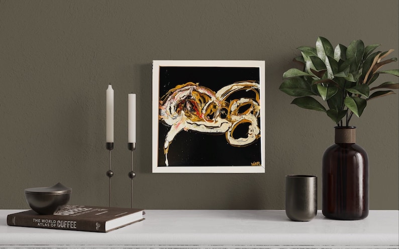 Puede incluir: Pintura abstracta en un marco blanco con pintura dorada, blanca y negra sobre fondo negro. La pintura est&aacute; colgada en una pared marr&oacute;n sobre una repisa blanca con un libro, velas y un jarr&oacute;n con hojas verdes.