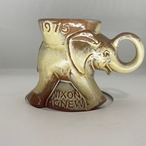 Puede incluir: Una taza de cerámica con forma de elefante con el texto "1973 Nixon Agnew" en la parte inferior. La taza es marrón y blanca con un acabado esmaltado.