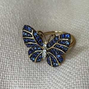 Könnte beinhalten: Ein goldfarbener Schmetterlingsring mit blauen Edelsteinen und einer kleinen Ansammlung weißer Edelsteine in der Mitte. Die Schmetterlingsflügel sind mit Reihen blauer Steine verziert. Der Ring ist vor einem neutralen Hintergrund platziert.