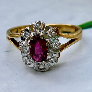 Peut inclure: Une bague en or avec une pierre précieuse rouge foncé de taille ovale au centre, entourée d'un halo de petites pierres claires de taille ronde serties dans une monture en métal argenté. L'anneau est en or jaune poli avec un design à double branche.