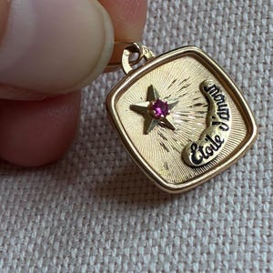 May include: A gold-toned pendant with a square frame. Inside, a star with a red gemstone sits above a curved banner with the word "Etoile". The pendant has a textured background with sunburst rays.