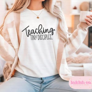 May include: White sweatshirt with the text "Teaching Tiny Disciples" in black script. The person is wearing a cream-colored cardigan with a striped pattern on the sleeves. A gold necklace is visible.