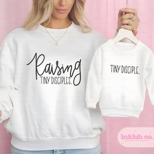 May include: White crewneck sweatshirt with the text "Raising Tiny Disciples."  A smaller white crewneck sweatshirt with the text "Tiny Disciple." is shown next to it.