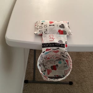 May include: A white fabric hanging organizer with a cylindrical pouch and a rectangular pouch. The organizer is attached to a table with a black metal frame. The pouches are decorated with a colorful print featuring knitting-themed words and images, including "I love to knit so much", "All you knit is joy", "Happy Place", and "I am a knitter".