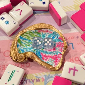 May include: A decorative oyster shell with a gold-painted rim and a colorful interior pattern of blue, green, and pink, containing two clear dice with white pips. The shell is surrounded by several white and pink mahjong tiles with various symbols.