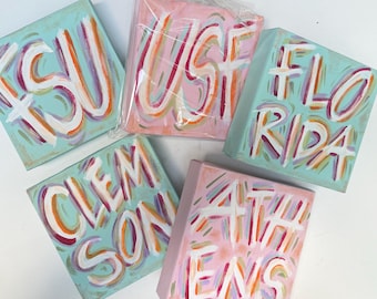 May include: Five square canvas paintings featuring hand-painted lettering in white with colourful outlines. The canvases display the text FSU, USF, FLORIDA, CLEMSON, and ATHENS. The backgrounds are light blue or pink, and one canvas is wrapped in plastic.